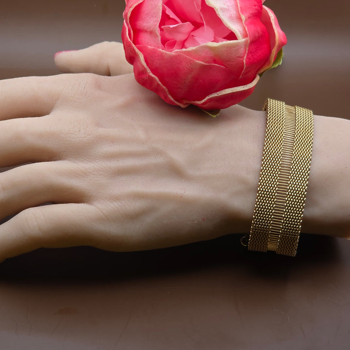 Hand wearing a gold bracelet with a pink flower against a brown background