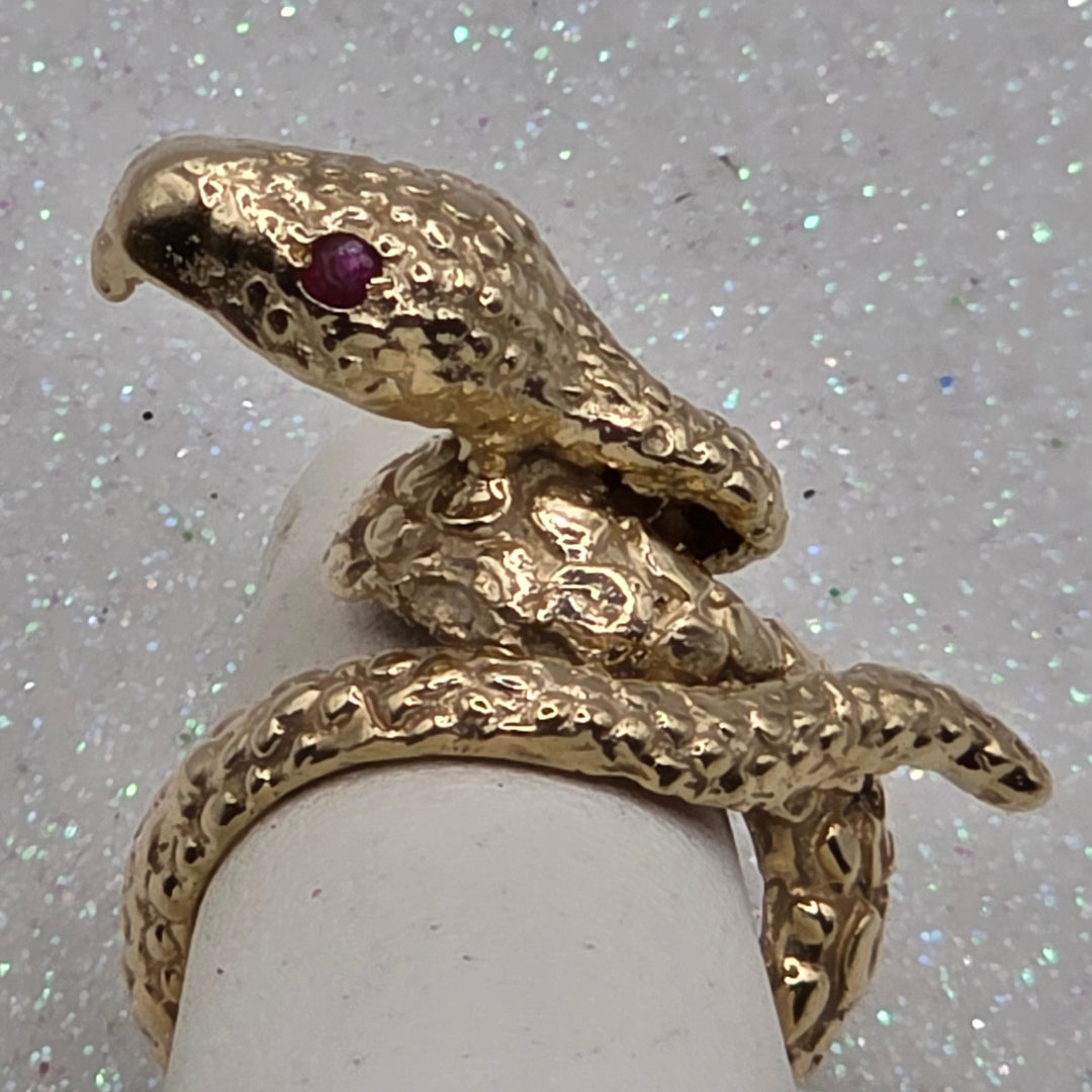 Gold snake-shaped ring with a red gemstone on a white stand against a glittery gray background